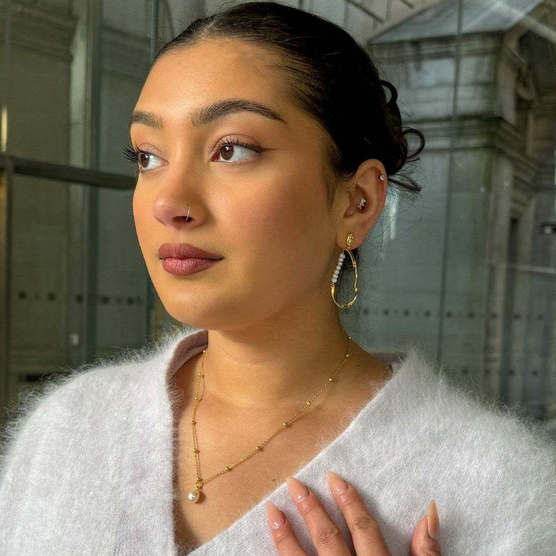 Woman wearing a light gray cardigan with gold buttons and jewelry, standing against a reflective glass wall.