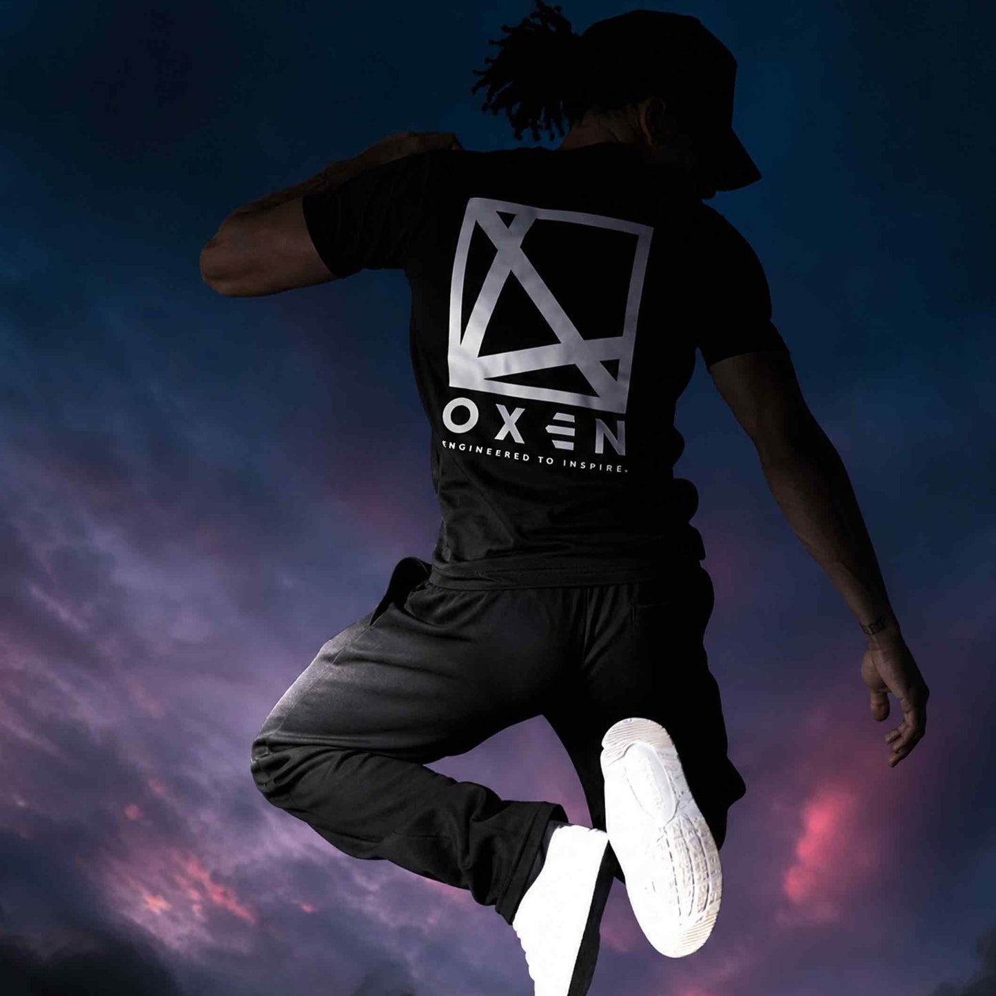 Person wearing a black t-shirt with a logo and text, jumping against a colorful sky.