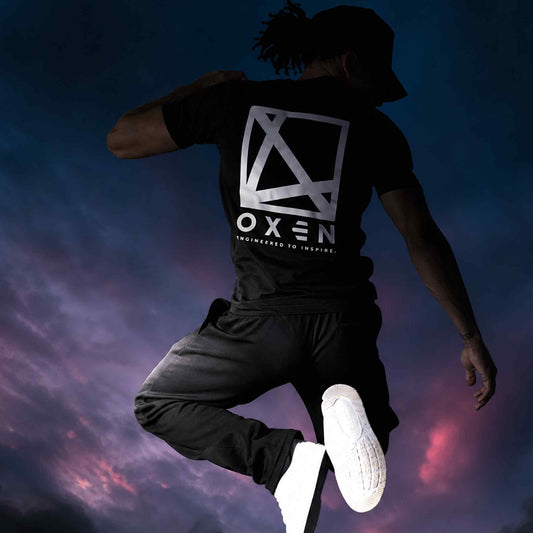 Person wearing a black t-shirt with a logo and text, jumping against a colorful sky.