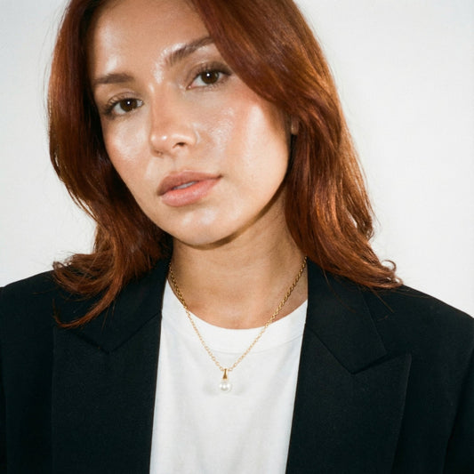 Woman wearing a black blazer and white shirt with a gold necklace against a plain background